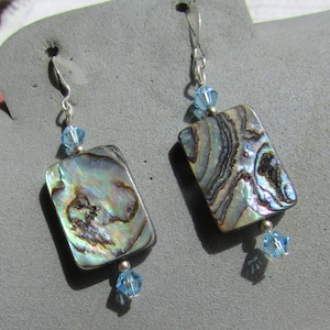 May include: A pair of rectangular abalone shell earrings with iridescent colors of blue, green, and brown. Each earring has a silver hook and is accented with a small blue crystal bead at the top and bottom.