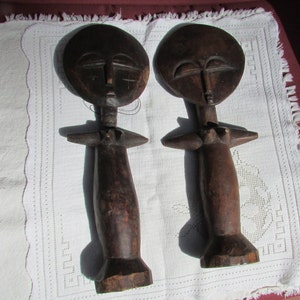 May include: Two wooden sculptures of stylized figures with round heads and simple features. The figures are dark brown and have a smooth, polished finish.