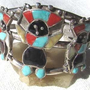 May include: Silver cuff bracelet featuring inlaid figures with turquoise, red, black, and white accents. The bracelet has a hinged design and is likely a piece of Native American jewelry.