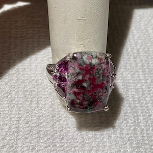 May include: A silver ring featuring a large, rectangular stone with a mottled pattern of pink, gray, and white. Smaller pink stones accent the sides of the ring, creating a striking contrast.