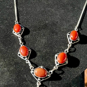 May include: A silver necklace and earring set featuring oval, reddish-orange gemstones set in diamond-shaped frames. The necklace has a pendant and the earrings have a hook closure. The jewelry is displayed on a black surface.