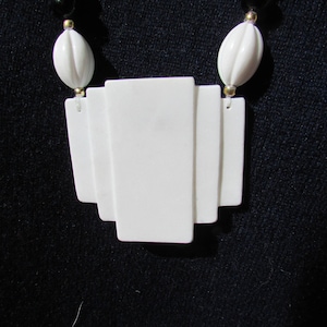 Puede incluir: Collar blanco de estilo Art Déco con colgante geométrico. El collar presenta un colgante rectangular en capas y cuentas ovaladas blancas. El collar tiene cuentas negras y detalles dorados. Es una pieza vintage.