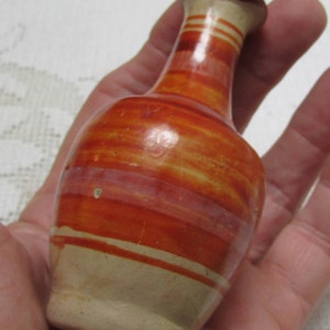 Puede incluir: Un pequeño jarrón de cerámica pintado a mano con un cuerpo bulboso y un cuello estrecho. El jarrón presenta bandas horizontales de colores naranja, rojo y crema. El jarrón mide aproximadamente 10 cm de alto.