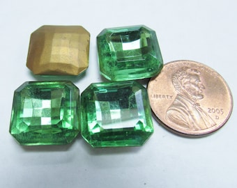 May include: An assortment of emerald-cut green glass gems and one gold-colored gem. A U.S. penny is included for size comparison. The gems are faceted and reflect light, showcasing their clarity and shape.