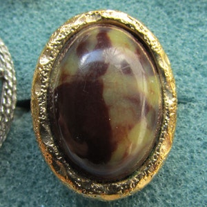 May include: Oval-shaped vintage earring with a gold-tone textured frame. The center features a polished stone with a mix of brown and green hues. The earring is set against a teal background.