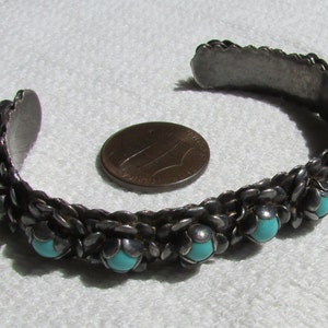May include: A silver bracelet with turquoise stones set in a woven design. The bracelet is about the size of a US penny.