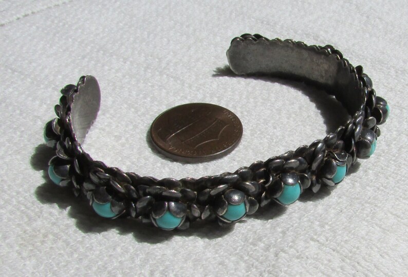 May include: A silver bracelet with turquoise stones set in a woven design. The bracelet is about the size of a US penny.