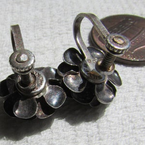 May include: A pair of silver tone flower shaped clip on earrings. Each earring has five petals and a screw back closure.