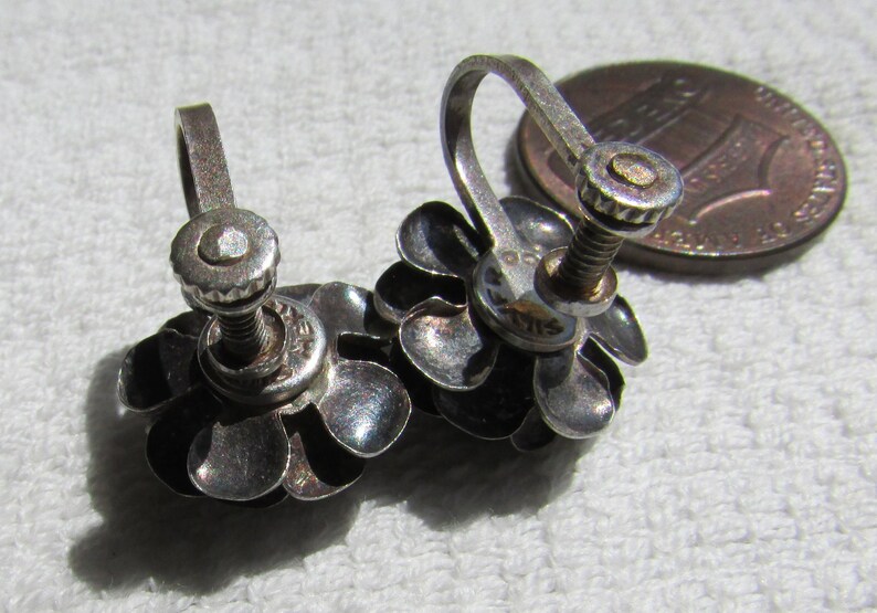 May include: A pair of silver tone flower shaped clip on earrings. Each earring has five petals and a screw back closure.