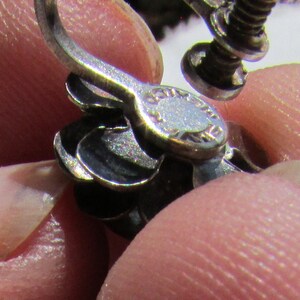 May include: Close-up of a silver earring with a screw-back closure. The earring has a decorative design with multiple layers.