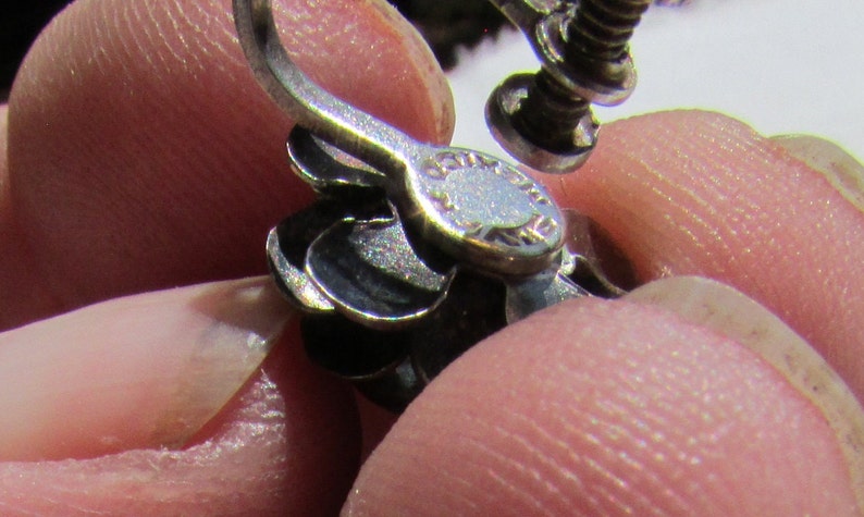 May include: Close-up of a silver earring with a screw-back closure. The earring has a decorative design with multiple layers.