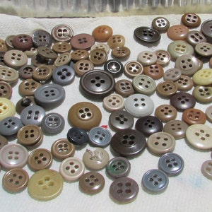 Puede incluir: Una colección de botones de varios tamaños en tonos marrones, beige y grises. Los botones, con cuatro agujeros, son de plástico. Están dispersos sobre una superficie blanca, ofreciendo una variedad para proyectos de costura y manualidades.