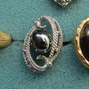May include: Silver-toned oval brooch with a dark, reflective center stone. The brooch features a textured, rope-like design around the stone, with a swirling, decorative shape. Other jewelry pieces are visible in the background.