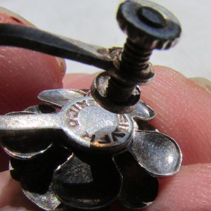 May include: Silver flower-shaped earring with a screw-back closure. The back of the earring has the words "925 STERLING" engraved on it.
