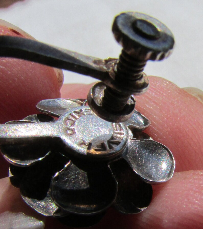 May include: Silver flower-shaped earring with a screw-back closure. The back of the earring has the words "925 STERLING" engraved on it.