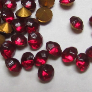 May include: A scattering of red, faceted, glass rhinestones with a gold metallic backing. The rhinestones are scattered on a white surface.