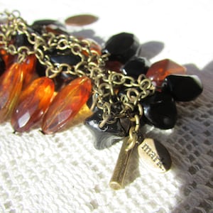 May include: A cluster of beads in shades of black and amber, strung on a gold-colored chain. The beads vary in shape, including faceted oblongs and rounded forms. A small charm with the word "mark" is attached.