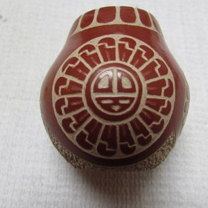 May include: A small, red clay pot with a white geometric design. The design features a stylized sun with a face and rays.