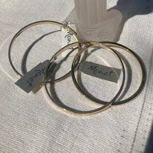 Puede incluir: Tres brazaletes de metal dorado con la marca "Monet" en etiquetas adjuntas. Los brazaletes circulares son de metal liso y brillante. Se encuentran sobre una superficie blanca y texturizada. Las etiquetas son grises con texto negro.