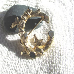 May include: A gold-toned brooch in the shape of a letter "C", crafted to resemble bamboo stalks and leaves. The brooch has a pin closure on the back. The design is detailed with textured surfaces and small, round accents.