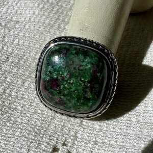 May include: A silver ring with a green and red gemstone set in a square bezel. The ring has a rope-like design around the edge of the bezel.