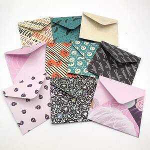 May include: A collection of colorful, folded paper envelopes. Some feature Halloween-themed patterns like skulls, bats, and pumpkins. Others have floral or geometric designs. The envelopes are in various sizes and colors, including pink, teal, and black.