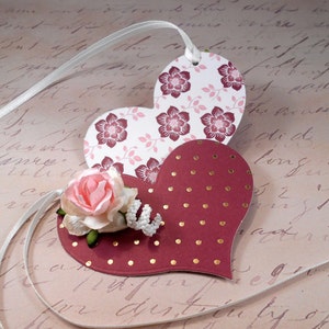 May include: Two heart-shaped paper tags with a white ribbon. The top tag is white with a pink and burgundy floral pattern. The bottom tag is burgundy with gold polka dots. A pink flower with green leaves is attached to the tags.