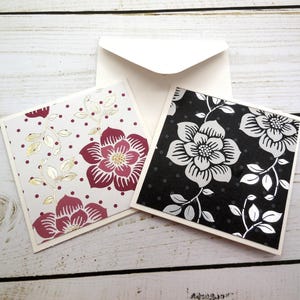 May include: Two blank greeting cards with floral patterns. One card has a white background with red flowers and gold accents. The other card has a black background with white flowers and silver accents.