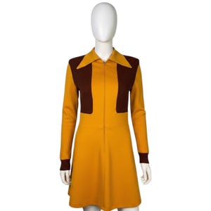 Puede incluir: Un vestido de manga larga con base de color amarillo mostaza, paneles acanalados de color marrón oscuro en el pecho y puños de color marrón oscuro. El vestido incluye un cuello puntiagudo y una cremallera vertical en la parte delantera.