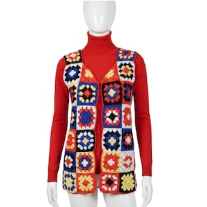 May include: A sleeveless crochet waistcoat featuring a multi-coloured granny square pattern in red, blue, yellow, white, and black. The waistcoat is worn over a solid red long-sleeved turtleneck jumper. The garment has a V-neckline and a straight hemline.
