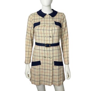 May include: A beige long-sleeve dress featuring a dark blue and red plaid pattern. The dress has a dark blue rounded collar, matching dark blue pocket flaps, and a double-breasted front with gold buttons. A dark blue belt with a gold buckle is included.