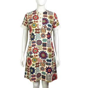 May include: A short-sleeved dress with a cream base color featuring a repeating pattern of red, blue, yellow, and green geometric shapes, sunbursts, and abstract symbols. The dress has a high neckline with a vertical placket containing four gold buttons.