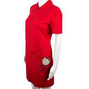 Puede incluir: Un vestido rojo brillante con mangas cortas y cierre de cremallera. El vestido presenta un cuello cosido en blanco y un bolsillo con un diseño de tulipán. El tulipán tiene un patrón de lunares blancos. Un vestido de inspiración vintage.
