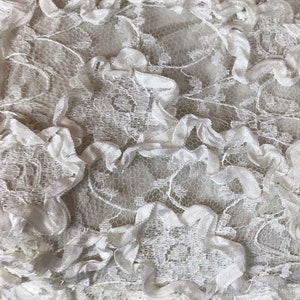 May include: Close-up of white lace fabric with a delicate floral pattern and ruffled trim. The lace has a textured appearance, with a combination of openwork and solid areas. The fabric is likely used for clothing or home decor.