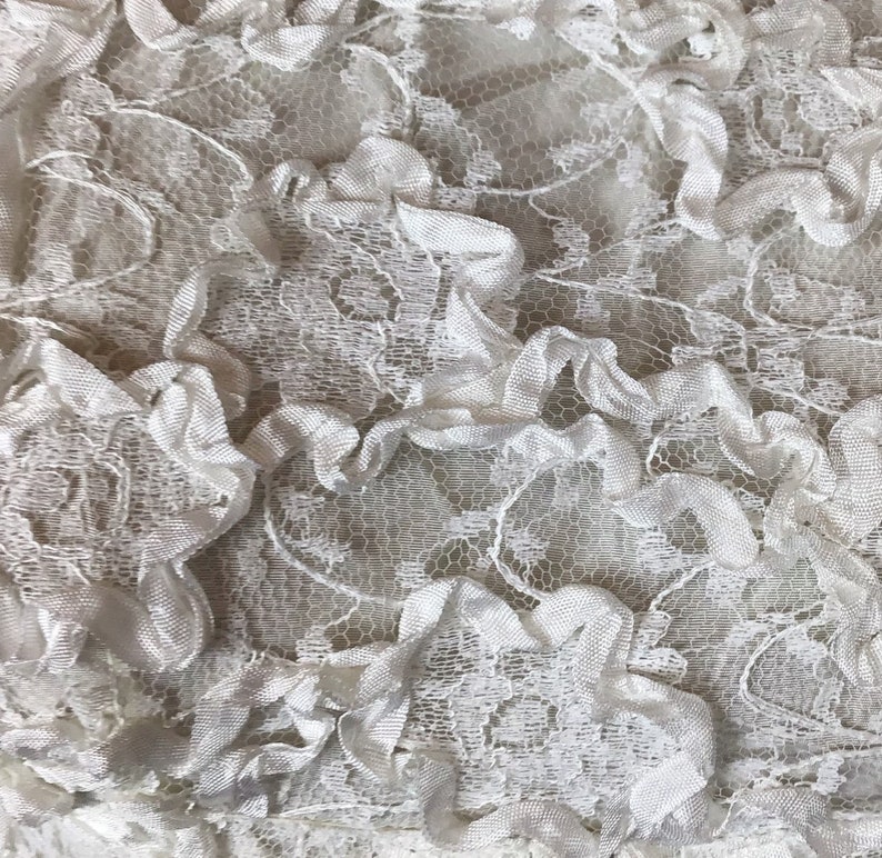 May include: Close-up of white lace fabric with a delicate floral pattern and ruffled trim. The lace has a textured appearance, with a combination of openwork and solid areas. The fabric is likely used for clothing or home decor.