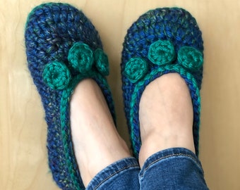Può includere: Un paio di pantofole all'uncinetto blu e verdi con pompon verdi sulla parte superiore. Le pantofole sono indossate da una persona che indossa jeans blu.