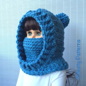 May include: A blue crocheted hooded cowl with a pom-pom on top. The cowl is worn by a person with brown hair and blue eyes.