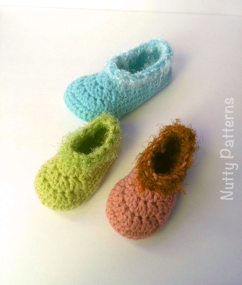May include: Three pairs of crocheted baby slippers in different colors: light blue, green, and pink. The slippers have a fluffy trim around the top.