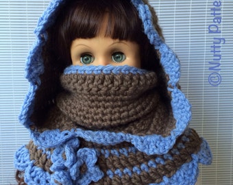 Può includere: Una sciarpa con cappuccio in crochet marrone e blu con un dettaglio floreale. La sciarpa è indossata da una bambola con i capelli castani e gli occhi azzurri.