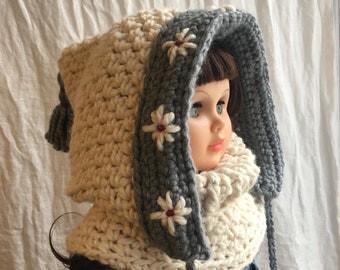 Può includere: Un cappello a uncinetto bianco e grigio con una sciarpa attaccata. Il cappello ha un cappuccio ed è decorato con fiori bianchi con centri rossi.