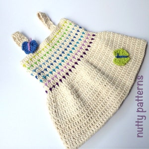 以下が含まれることがあります： 虹色のストライプ模様と蝶の装飾が施された、かぎ針編みのベビーワンピース。ワンピースはベージュの糸で編まれており、シンプルでクラシックなデザインです。