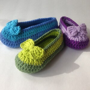 May include: Three pairs of crocheted baby shoes in different colors. The shoes are made with a striped pattern and have a bow on the top. The colors are blue, green, and purple.