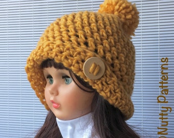 Può includere: Un berretto di lana giallo con un pon pon in cima e un bottone di legno. Il berretto è indossato da una bambola con i capelli castani.