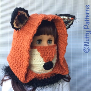 May include: A crocheted fox hood with a white face and black nose. The hood is orange and has a black and white trim. The hood is worn by a doll.