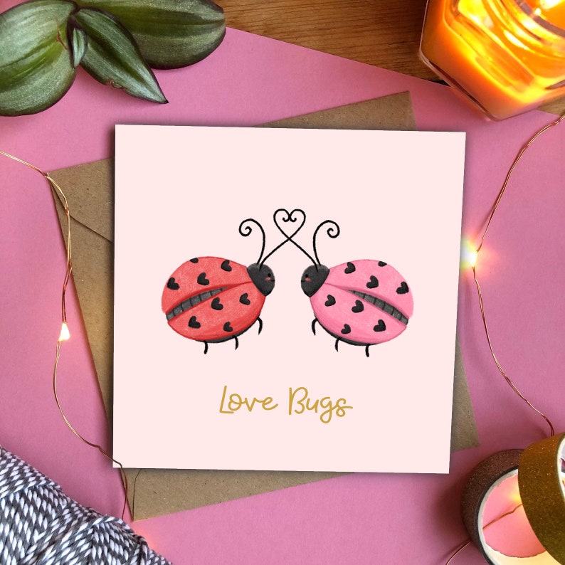 Love Bugs Valentines Card depicts two ladybugs ladybirds with hearts instead of spots looking lovingly at each other. One is pink & one is red. Their antenna are intertwined to make a heart. Text below reads love bugs in gold script writing.