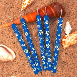 May include: A collection of five blue quilling tools with white and yellow floral patterns. The tools are arranged on a sandy surface, surrounded by seashells in shades of orange, white, and brown.