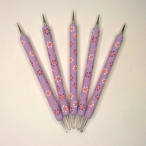 May include: Five lavender dotting tools with silver tips, arranged in a fan shape. The handles are decorated with a repeating pattern of white, orange, and purple floral designs. These tools are used for nail art and crafts.