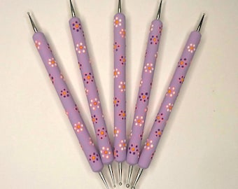 Puede incluir: Cinco herramientas de punteado lavanda con puntas plateadas, dispuestas en forma de abanico. Los mangos están decorados con un patrón floral repetido en blanco, naranja y morado. Estas herramientas se utilizan para nail art y manualidades.