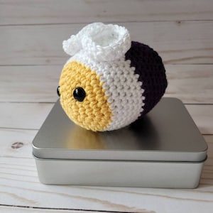 May include: A crocheted plush toy shaped like a bee with yellow, white, and purple stripes. The bee has a white crocheted crown on its head and black button eyes.