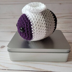 May include: A crocheted plush toy in the shape of a bee with a purple, gray, and white striped body and black eyes. The bee is sitting on a silver tin.